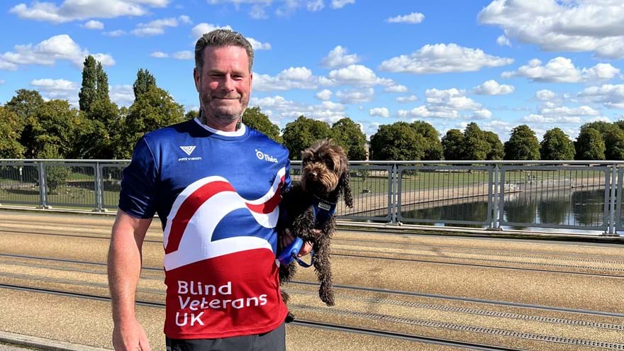 Simon stood in front of a canal in the sunshine wearing his Blind Veterans UK top and holding his dog