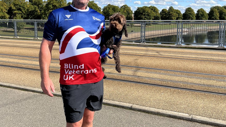 Simon stood in front of a canal in the sunshine wearing his Blind Veterans UK top and holding his dog