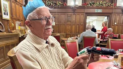 A man wearing a cream cardigan and a blue paper hat holds a blue christmas cracker