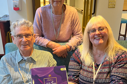 Three people sitting on a sofa, holding a box of Cadbury's milk tray chocolate and smiling