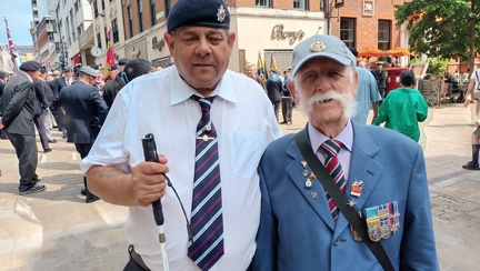 Chris and Michael are stood beside each other. Chris has his white cane in his hand and Michael is wearing a suit jacket and displaying his medals