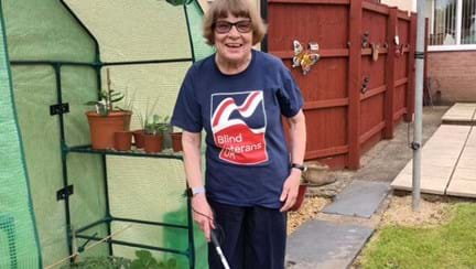 Noeline wearing Blind Veterans UK t-shirt stood in her garden holding her cane