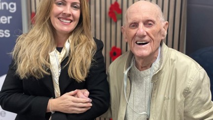 Kelly and Les both smiling are stood hand in hand in front of a display of poppies on the wall at the Centre of Wellbeing in Llandudno