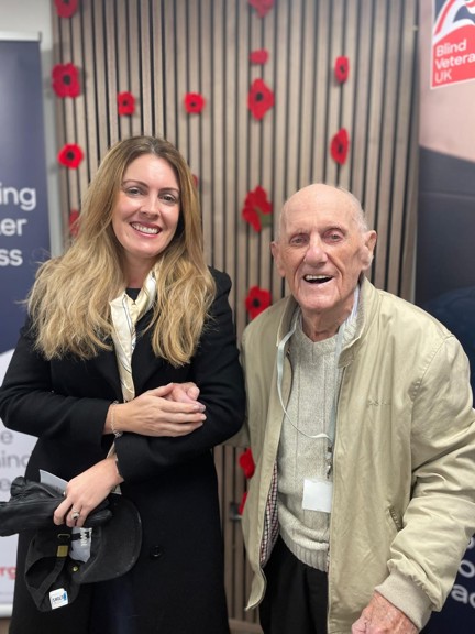 Kelly and Les both smiling are stood hand in hand in front of a display of poppies on the wall at the Centre of Wellbeing in Llandudno