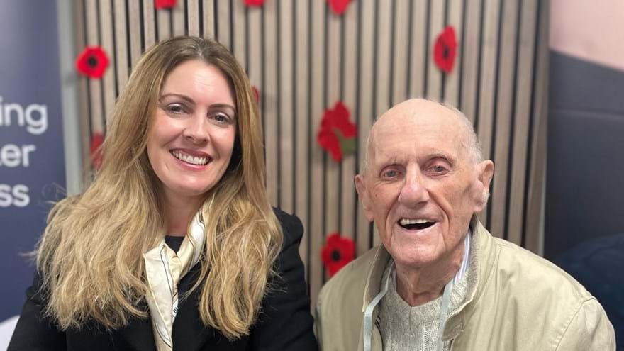 Kelly and Les both smiling are stood hand in hand in front of a display of poppies on the wall at the Centre of Wellbeing in Llandudno