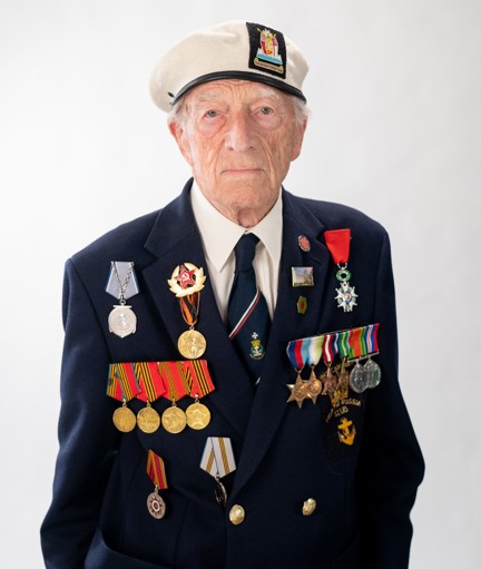 Alec is wearing a navy blazer, beret, medals, and looks into the camera proudly