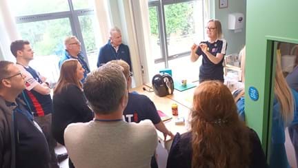 Blind Veterans UK Occupational Therapist Catherine in the Llandudno Centre of Wellbeing training kitchen with a group of Thea team members, demonstrating equipment provided to our blind veterans to help them live with sight loss