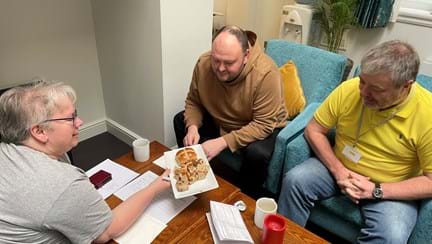 A lady holds a plate of hot cross buns to two men sat opposite her at a coffee table