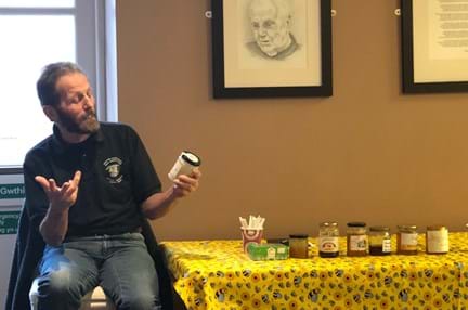 A beekeeper sat next to a table with a yellow bee-themed tablecloth, holding a jar of honey and giving a talk