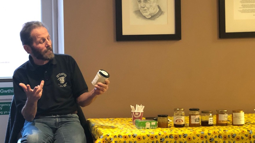 A beekeeper sat next to a table with a yellow bee-themed tablecloth, holding a jar of honey and giving a talk