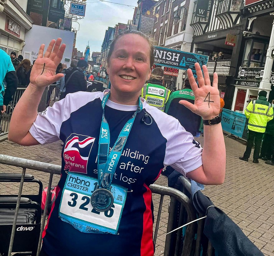 Supporter Alison wearing her marathon medal and Blind Veterans UK training top, holding up her hands to show her palms, with the number 1 written on her left palm, and 4 on her right palm.