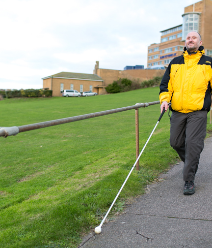 Blind veteran Mark using a guide cane to walk down a path in front of Blind Veterans UK centre of wellbeing