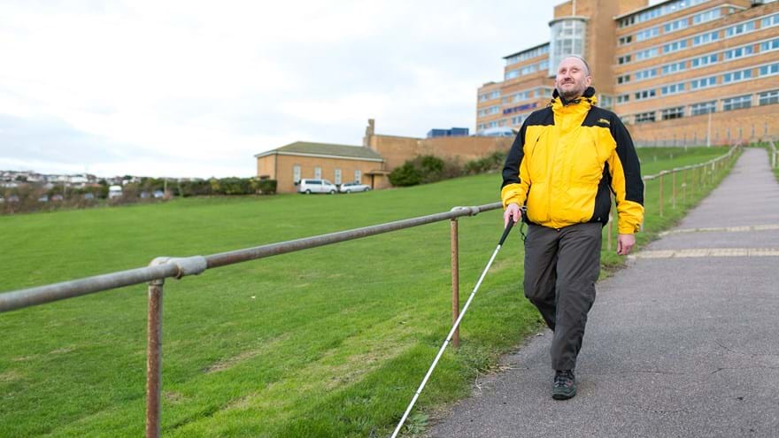 Blind veteran Mark using a guide cane to walk down a path in front of Blind Veterans UK centre of wellbeing