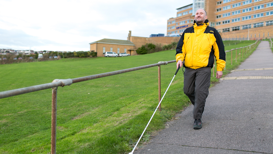 Blind veteran Mark using a guide cane to walk down a path in front of Blind Veterans UK centre of wellbeing