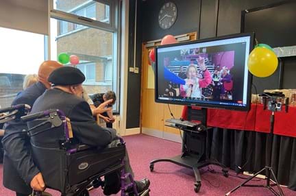 Blind veteran Ken in front of a TV screen with Margaret waving from the video call