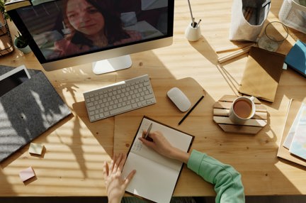 A woman sat at a desk writing in a notebook while on a computer video call