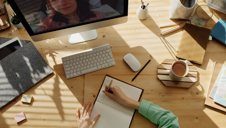 A woman sat at a desk writing in a notebook while on a computer video call