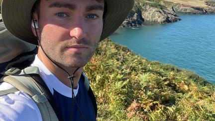 James wearing a Blind Veterans UK t shirt stood on the coastal path on a very sunny day with the sea in the background