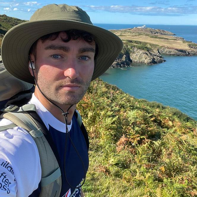 James wearing a Blind Veterans UK t shirt stood on the coastal path on a very sunny day with the sea in the background