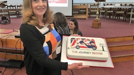 Jennie looks into camera and is holding a cake with a bus on it in the Blind Veterans UK colours. The caption below the bus reads 'the journey begins'.