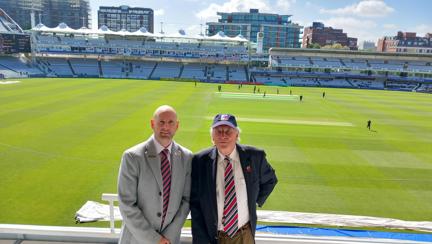 Chris and Tim stood side by side with play taking place on the cricket pitch behind them