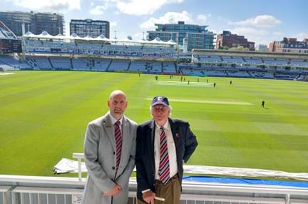 Chris and Tim stood side by side with play taking place on the cricket pitch behind them