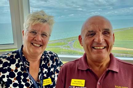 A photo of our Centre Manager, Lesley, left, with blind veteran Alan, right. Both smiling and looking at camera with background overlooking the sea.