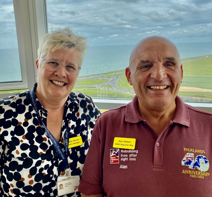 A photo of our Centre Manager, Lesley, left, with blind veteran Alan, right. Both smiling and looking at camera with background overlooking the sea.