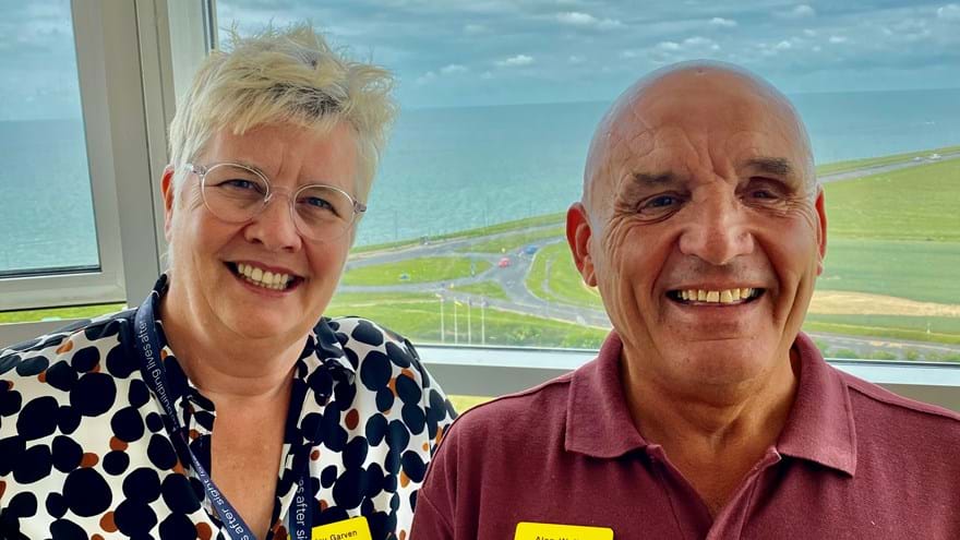 A photo of our Centre Manager, Lesley, left, with blind veteran Alan, right. Both smiling and looking at camera with background overlooking the sea.