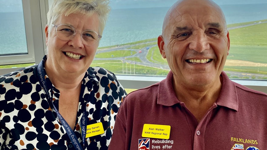 A photo of our Centre Manager, Lesley, left, with blind veteran Alan, right. Both smiling and looking at camera with background overlooking the sea.