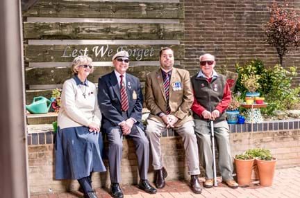A group of blind veterans seated on a wall, smiling in front of a sign that reads "Lest We Forget"