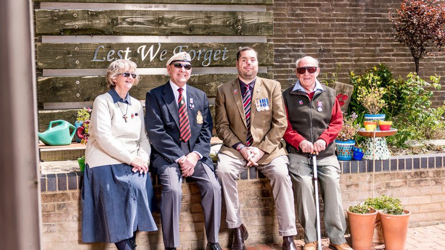 A group of blind veterans seated on a wall, smiling in front of a sign that reads "Lest We Forget"