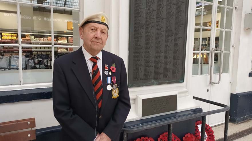 Tony wearing his medals and stood in front of Brighton Train Station War Memorial which has a number of poppy wreaths laid against it