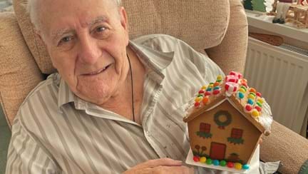 Blind veteran Maurice looks into the camera and smiles while holding a small gingerbread house