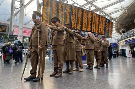 The men line up behind each other with their hands on each other's shoulders, train schedule screens are in the background