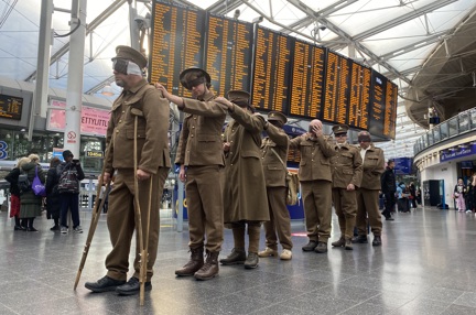 The men line up behind each other with their hands on each other's shoulders, train schedule screens are in the background