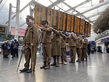 The men line up behind each other with their hands on each other's shoulders, train schedule screens are in the background