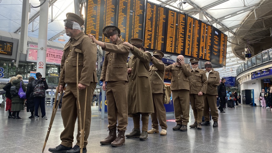 The men line up behind each other with their hands on each other's shoulders, train schedule screens are in the background