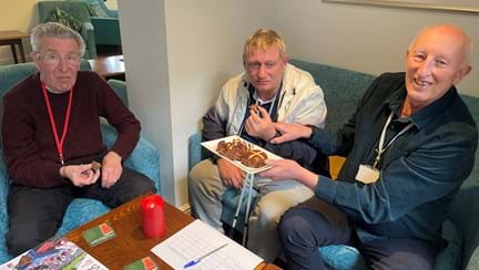 Three men sit at a table, two are holding pieces of hot cross bun, while the third holds a plate of hot cross bun pieces.