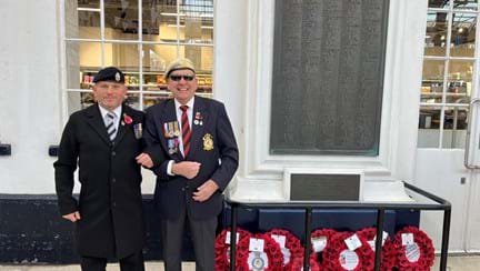 Blind veteran Tony with Brighton station manager Danny standing next to the Brighton station memorial with poppy wreaths at the bottom