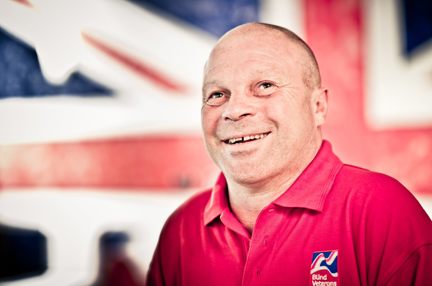 Blind veteran Billy smiling, wearing a red Blind Veterans UK polo shirt, with a British flag design covering the wall behind him.