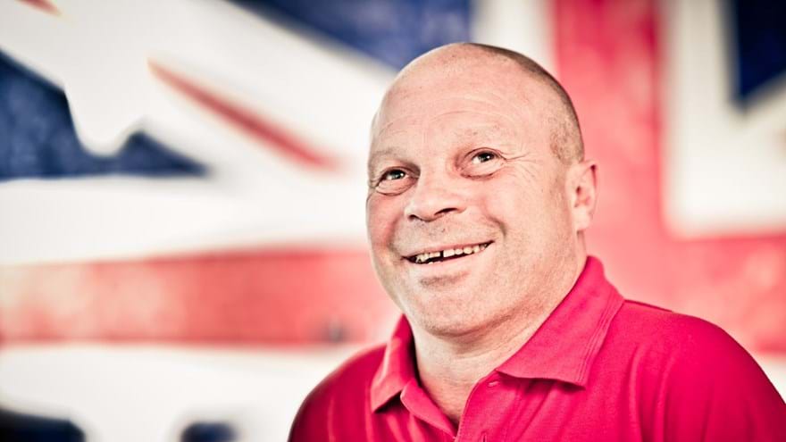 Blind veteran Billy smiling, wearing a red Blind Veterans UK polo shirt, with a British flag design covering the wall behind him.