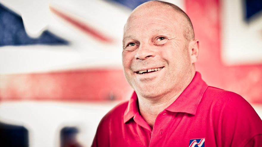 Blind veteran Billy smiling, wearing a red Blind Veterans UK polo shirt, with a British flag design covering the wall behind him.