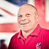 Blind veteran Billy smiling, wearing a red Blind Veterans UK polo shirt, with a British flag design covering the wall behind him.