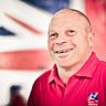 Blind veteran Billy smiling, wearing a red Blind Veterans UK polo shirt, with a British flag design covering the wall behind him.