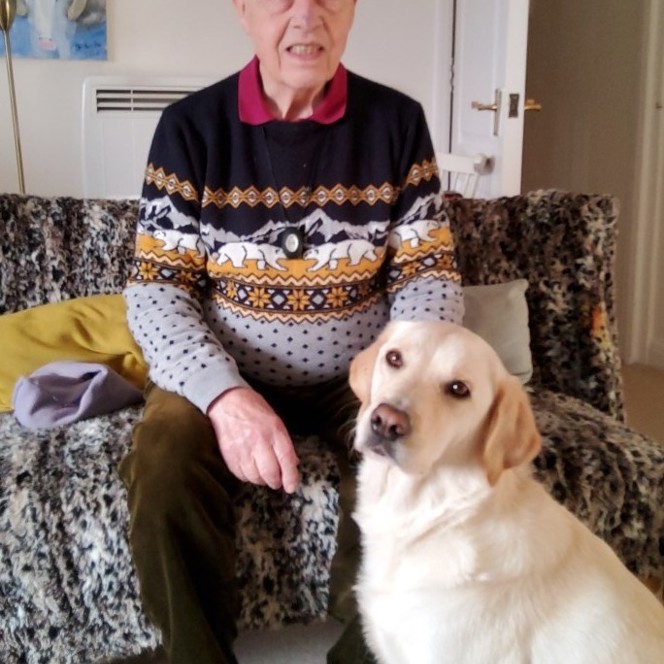 Tom is wearing a Christmas jumper and is sitting on the sofa with his guide dog at his feet