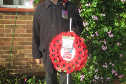 Blind Veteran Jim stood in his garden wearing a poppy and displaying his medals. He is holding his white cane and his poppy wreath.