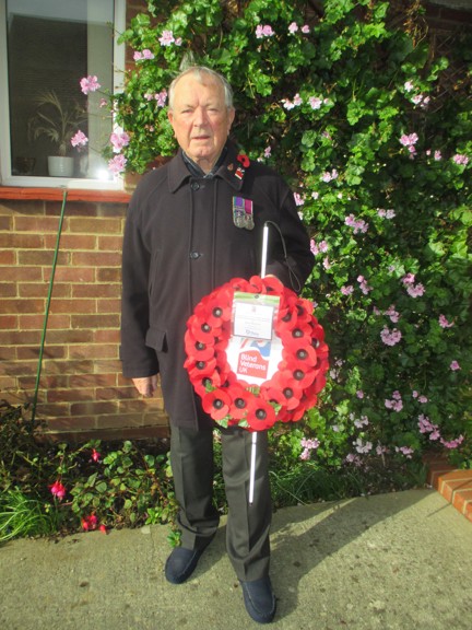Blind Veteran Jim stood in his garden wearing a poppy and displaying his medals. He is holding his white cane and his poppy wreath.