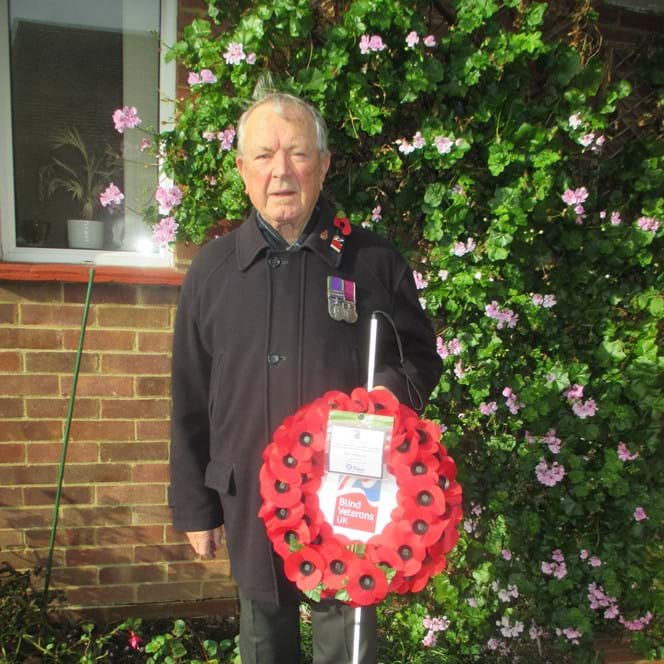 Blind Veteran Jim stood in his garden wearing a poppy and displaying his medals. He is holding his white cane and his poppy wreath.