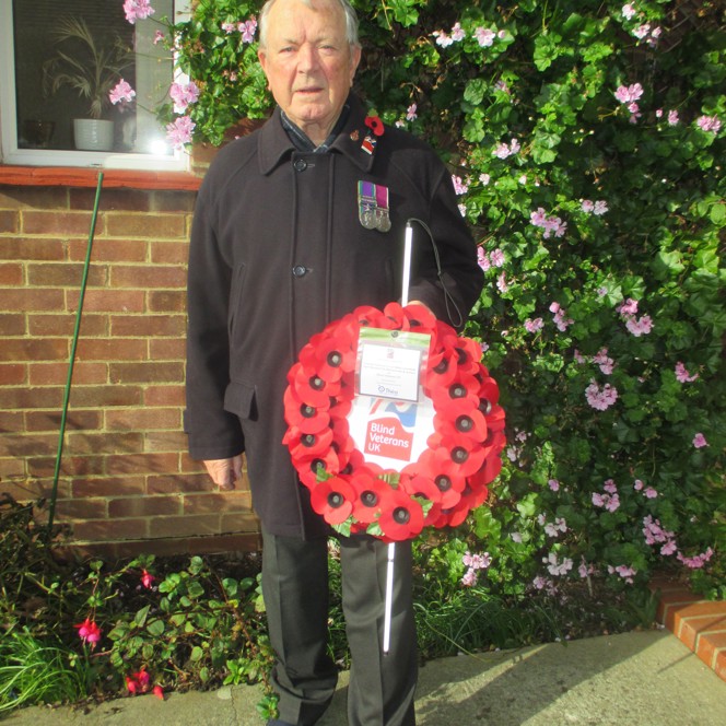 Blind Veteran Jim stood in his garden wearing a poppy and displaying his medals. He is holding his white cane and his poppy wreath.